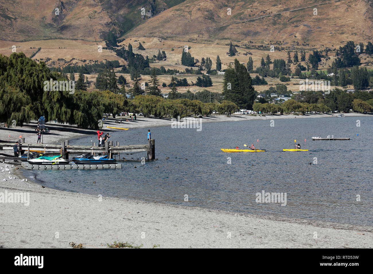 Persone in giallo kayak sul lago Wanaka dal jetty di turisti e di jet sci su una soleggiata giornata estiva, Wanaka, Nuova Zelanda Isola del Sud Foto Stock