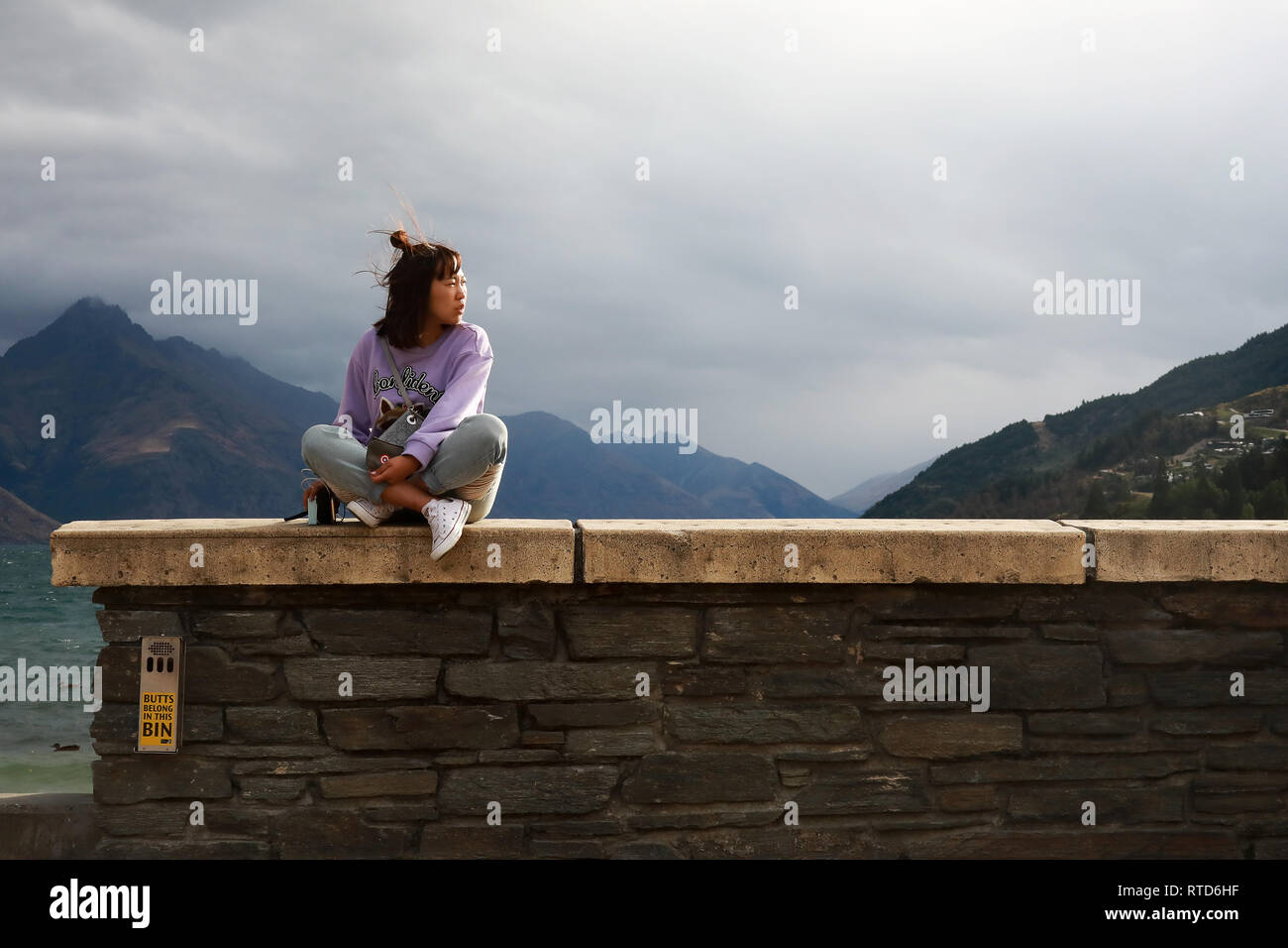 Asian tourist tenendo un sacchetto si siede sulla spiaggia parete in Queenstown affacciato sul lago Wakatipu'Isola Sud della Nuova Zelanda Foto Stock