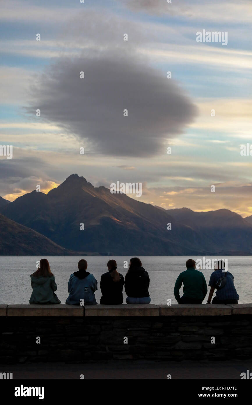 Turisti che si siedono sulla parete in spiaggia a guardare il tramonto sul lago Wakatipu, Queenstown, New Zealand Nuova Zelanda Isola del Sud Foto Stock