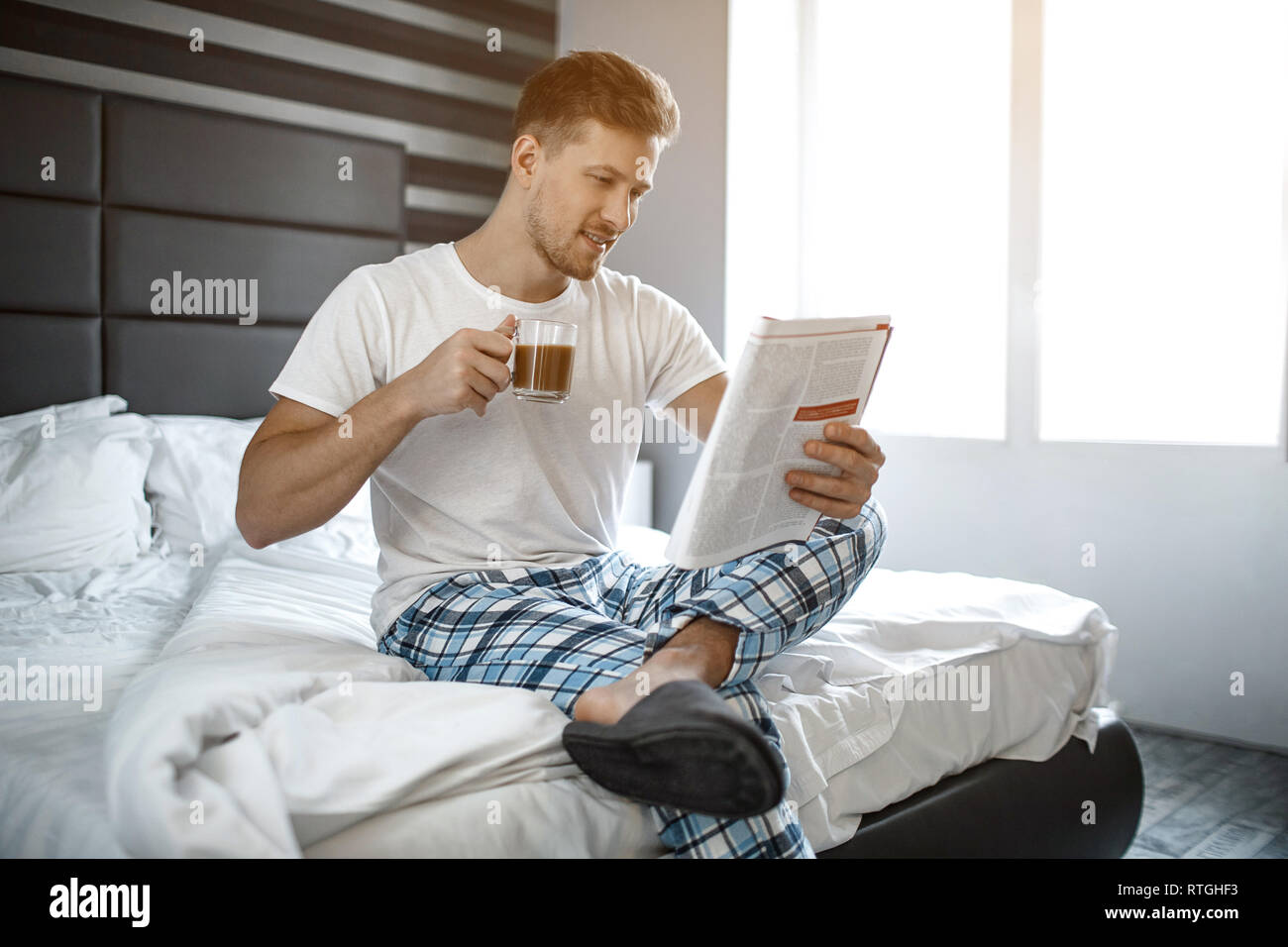 Giovane uomo sul letto la mattina. Egli sedersi e leggere ufficiale Guy tenere tazza di caffè. Piacevole e allegro Foto Stock