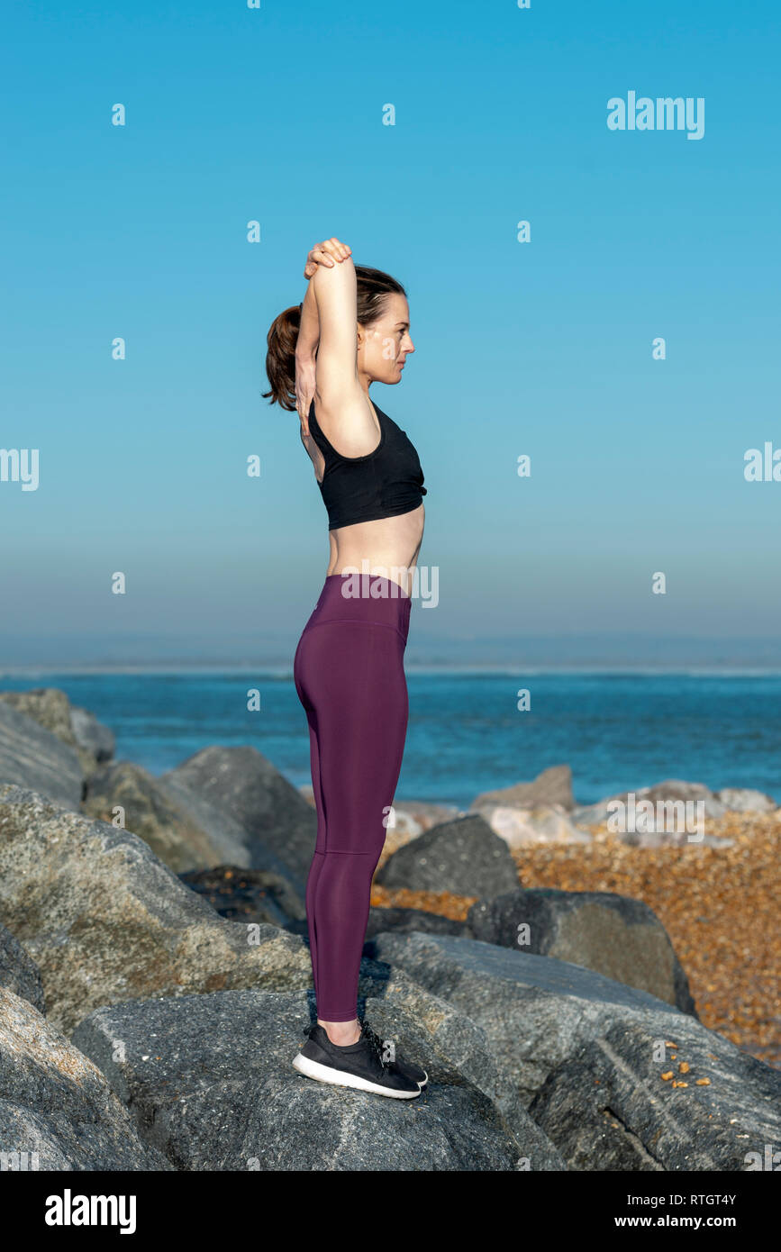 Donna facendo un tratto del braccio esercizio di riscaldamento esterno su rocce con cielo blu Foto Stock