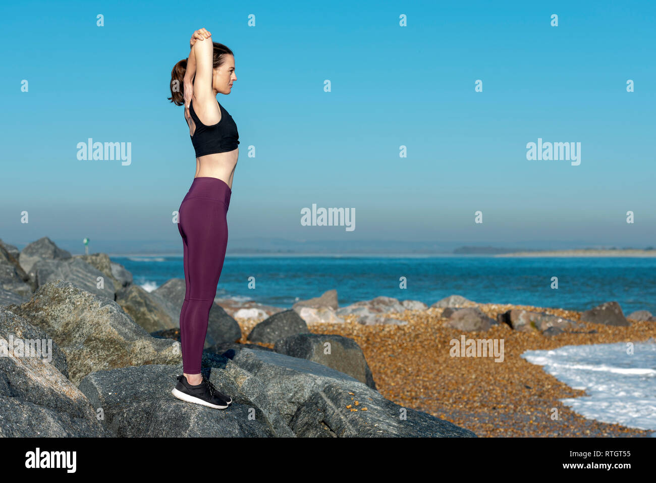 Donna facendo un tratto del braccio esercizio di riscaldamento esterno su rocce con cielo blu Foto Stock