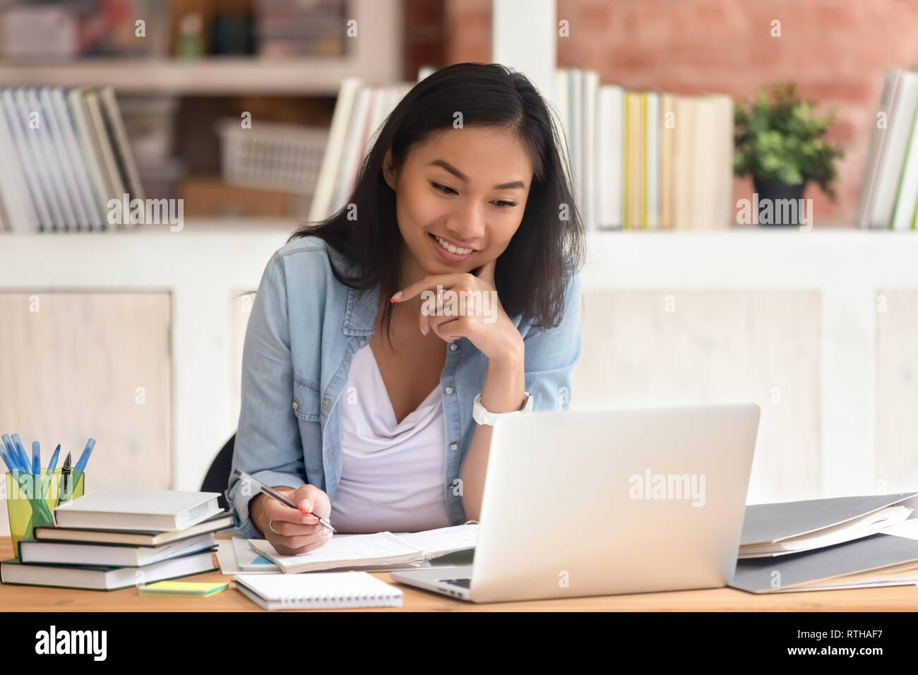 Sorridente ragazza asiatica studente studiare in biblioteca con libri per laptop Foto Stock