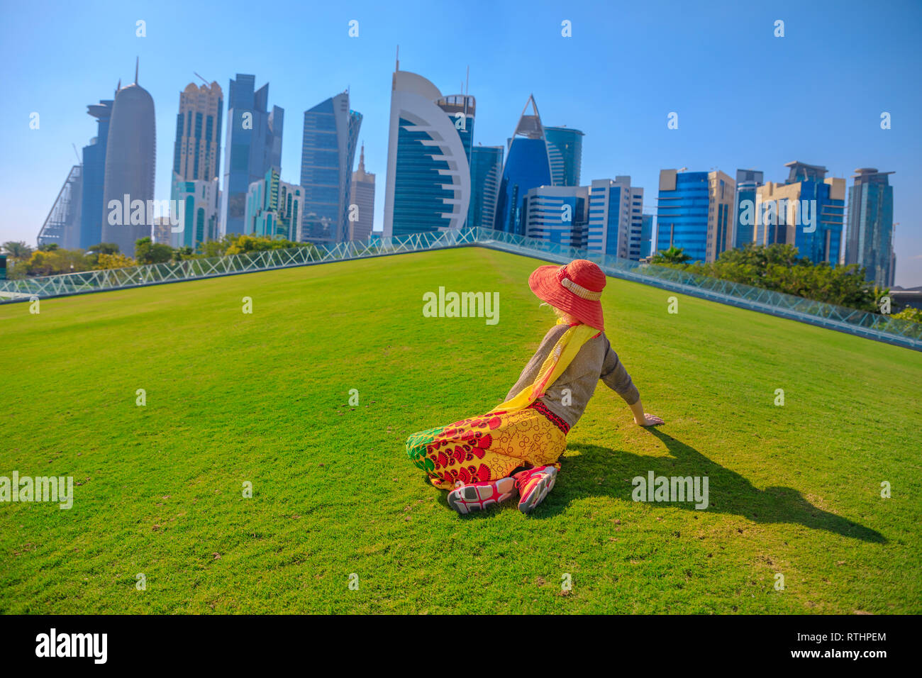 Viaggiatori di donna con cappello seduti su un prato verde in un parco con grattacieli di West Bay in background. Turista femminile gode di Doha skyline del centro Foto Stock