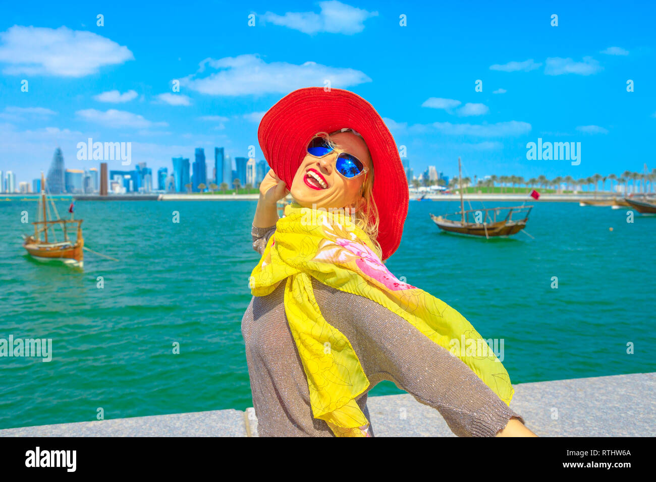 Felice donna con cappello lungo la baia di Doha con un tradizionale vecchio barche dhow su sfondo. Stile di vita turistica caucasica gode di grattacieli di Doha Foto Stock