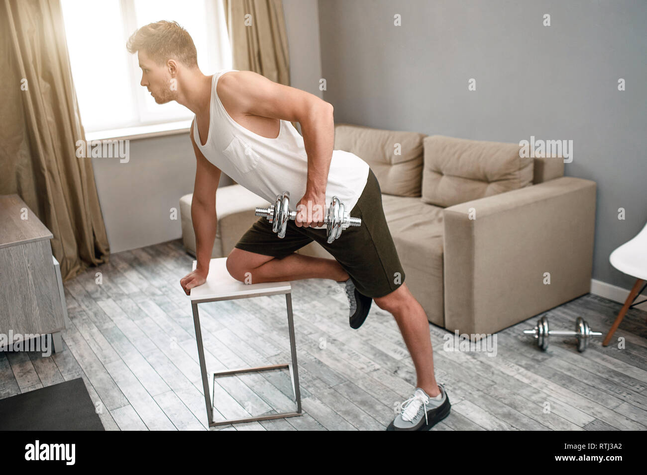 I giovani ben costruito uomo praticare sport in appartamento. Egli fa dumbbell esercizio di riga. Forte concentrato muscoloso giovane uomo Foto Stock