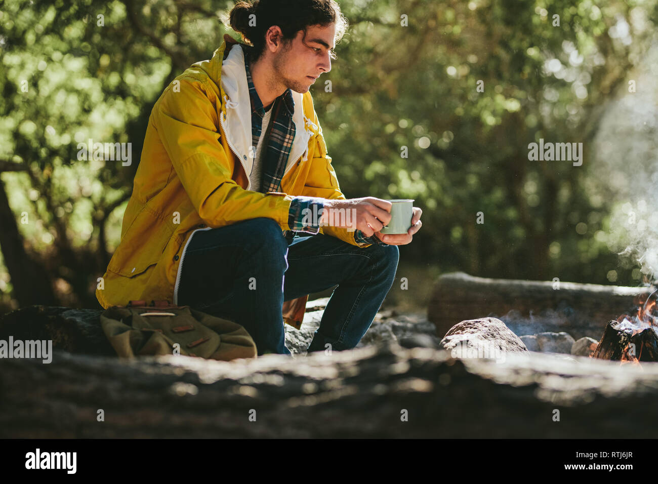 Vista laterale di un uomo seduto accanto a un falò in possesso di una tazza da caffè. Giovane uomo nel profondo pensiero guardando il fuoco. Foto Stock