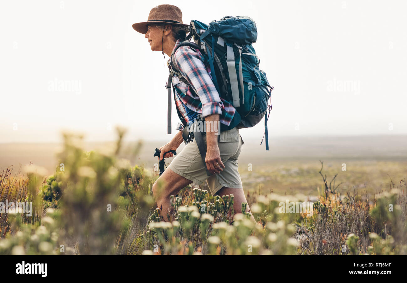Vista laterale di un senior Woman Trekking nel selvaggio. Donna che indossa uno zaino e hat a piedi attraverso cespugli e piante durante trekking tenendo un escursionismo po Foto Stock