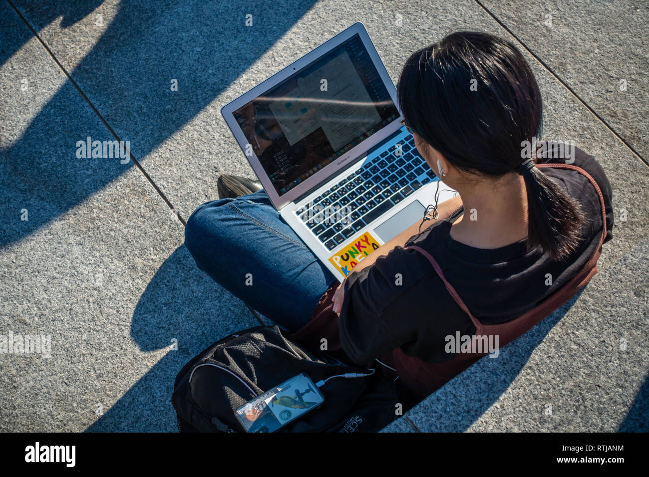 Immagine da sopra della giovane donna seduta al di fuori nel sole e utilizzando il suo laptop e telefoni cellulari a gocce di carbone cantiere, Kings Cross a Londra, Inghilterra Foto Stock