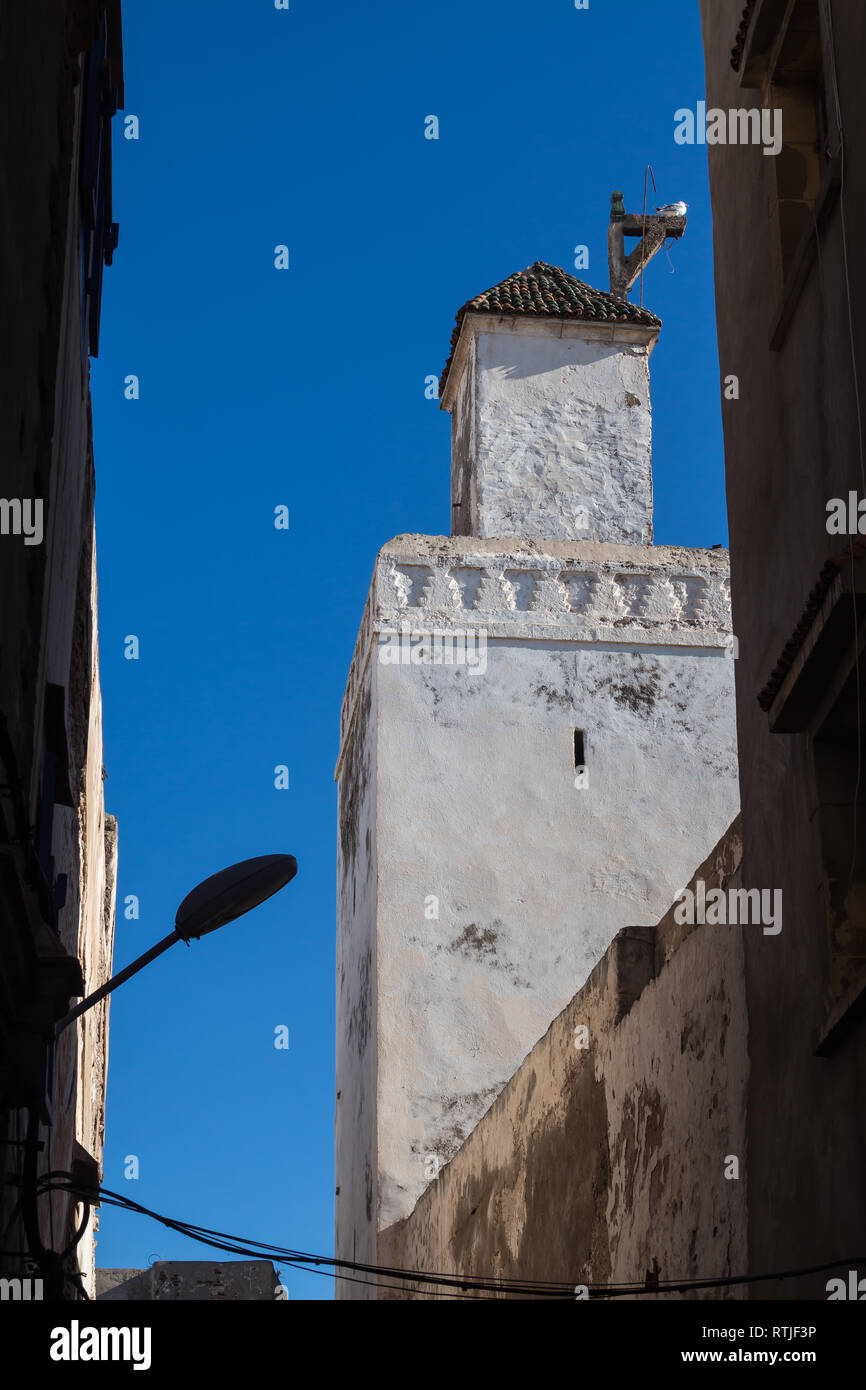 Torre di una moschea con la vecchia facciata bianca. Edifici del hosues intorno a. Silhouette di un lampione. Azzurro cielo. Essaouira, Marocco. Foto Stock