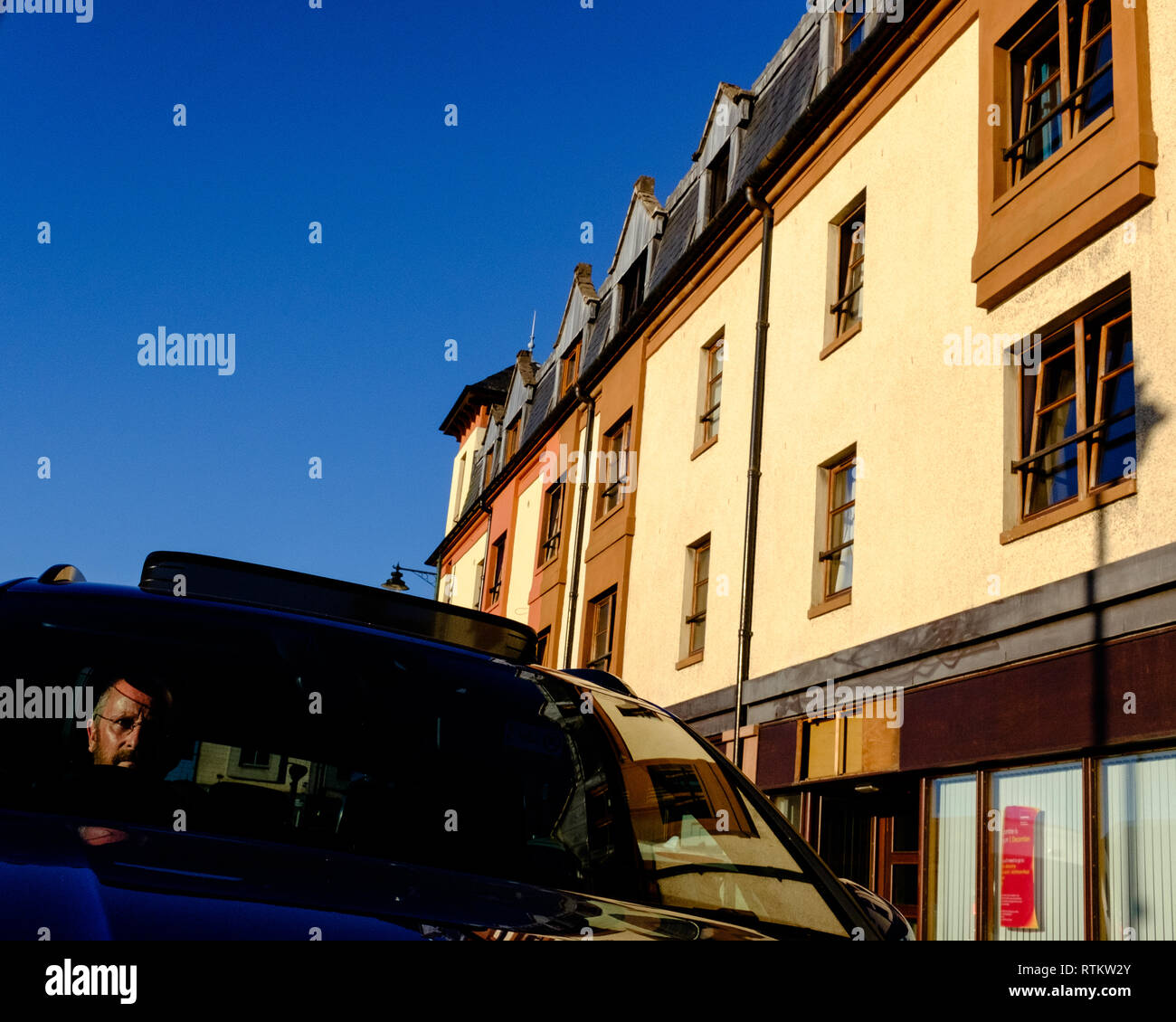 Uomo seduto in macchina con lo sguardo dritto con un giorno di bluebird e gli edifici residenziali che lo circonda. Fort William highlands scozzesi uk Foto Stock