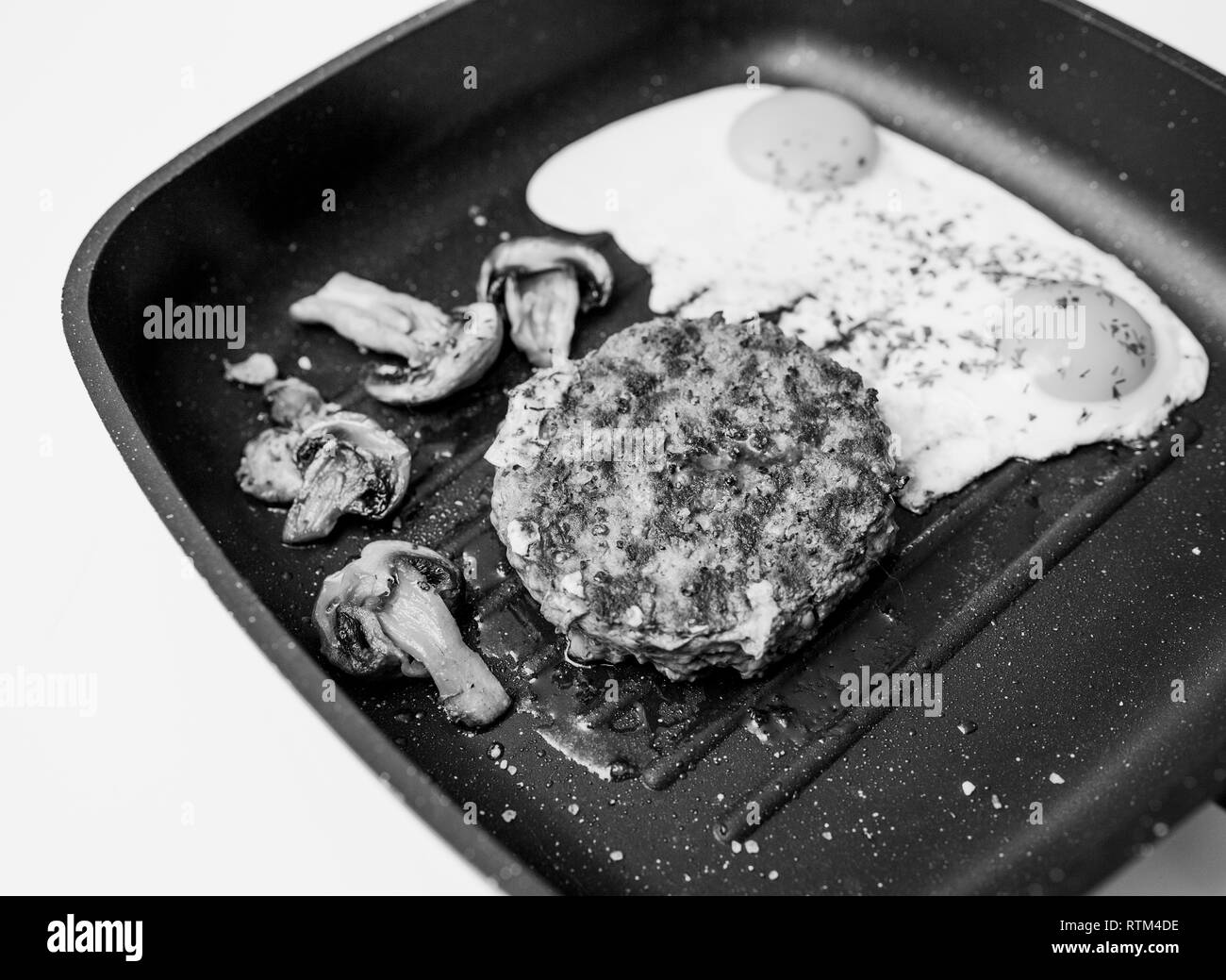 Al di sopra della deliziosa carne di manzo crudo hamburger di carne cotta in padella su teflon grill superficie in cucina con due uova fritte con organici champignon fritti immagine in bianco e nero Foto Stock