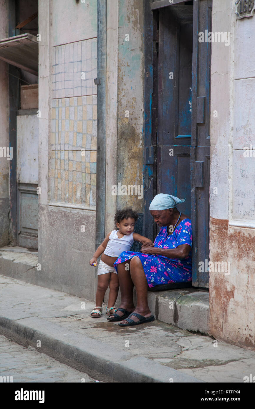 L'Avana, Cuba - 21 Gennaio 2013: una vista delle strade della città con il popolo cubano. Una nonnina con il bambino seduto sul gradino della porta. Foto Stock