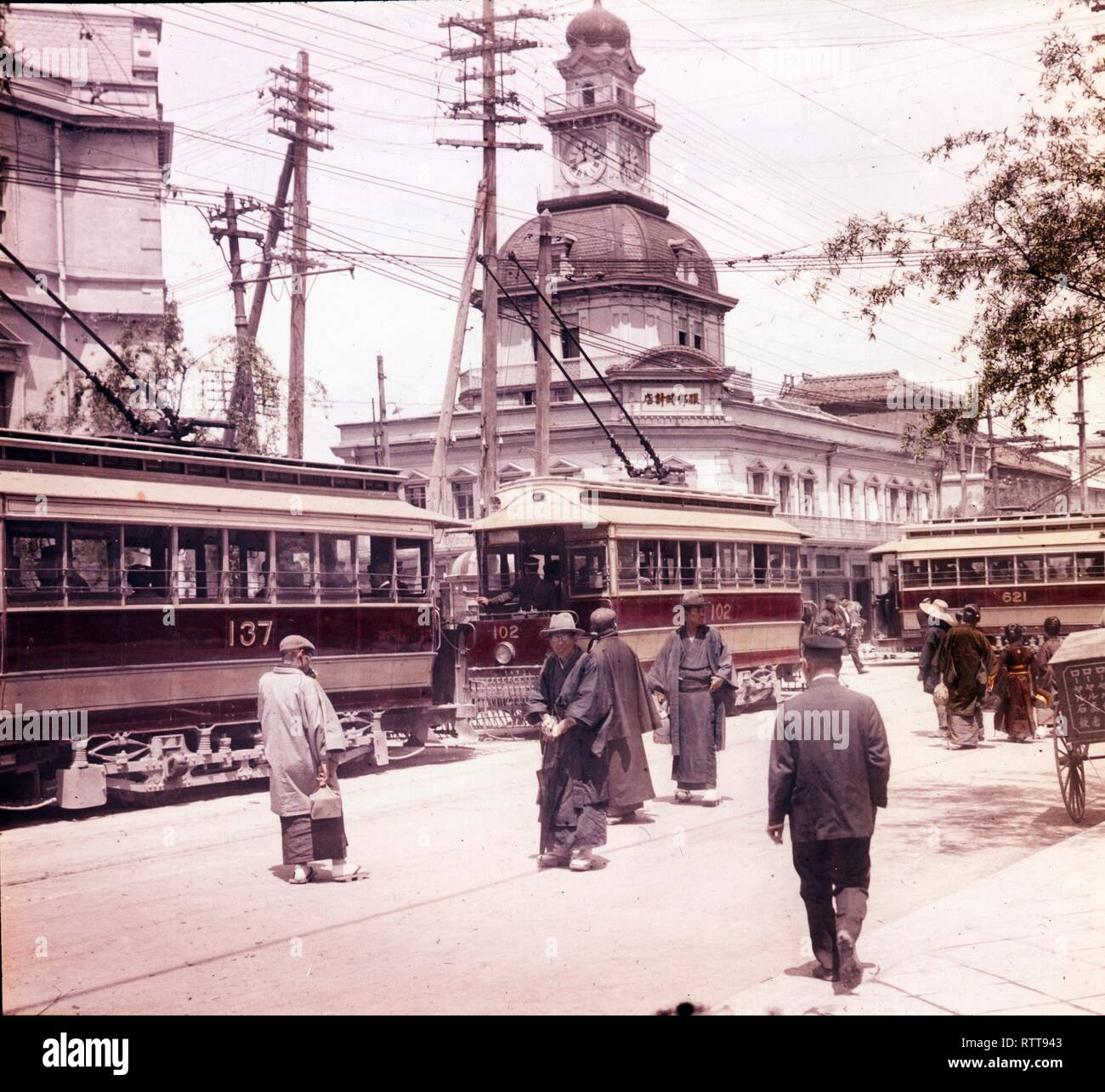 Foto colorata di pedoni e tram sul Ginza Crossing con l'edificio Hattori e la torre dell'orologio dall'altra parte della strada, Tokyo, Giappone, all'inizio degli anni '20 circa. (Foto di Burton Holmes) Foto Stock
