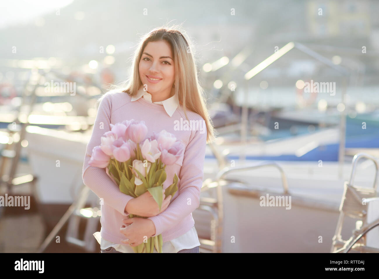 Bella ragazza in primavera o in autunno, in riva al mare nella baia di barca. Con un bouquet di fiori nelle sue mani e serrati eleganti jeans e un maglione rosa Foto Stock