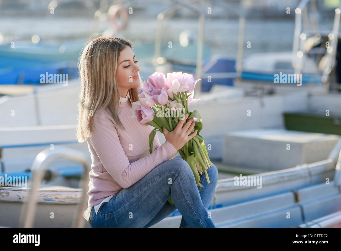 Bella ragazza in primavera o in autunno, in riva al mare nella baia di barca. Con un bouquet di fiori nelle sue mani e serrati eleganti jeans e un maglione rosa Foto Stock