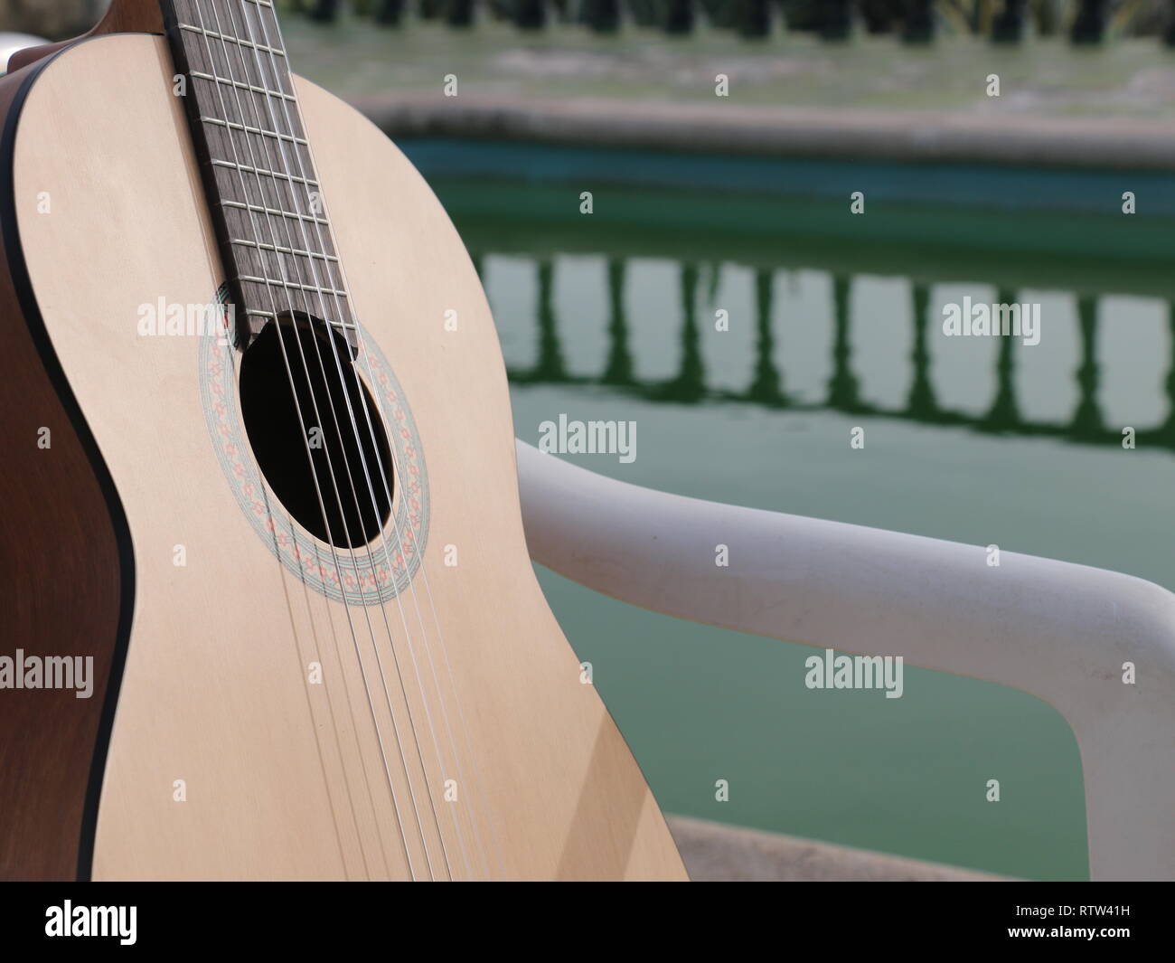 Un creativo il concetto di cattura di un legno lucido di chitarra acustica con un background palmtree e di una piscina a specchio di acqua e un pavimento verde dietro alla luce del sole. Foto Stock