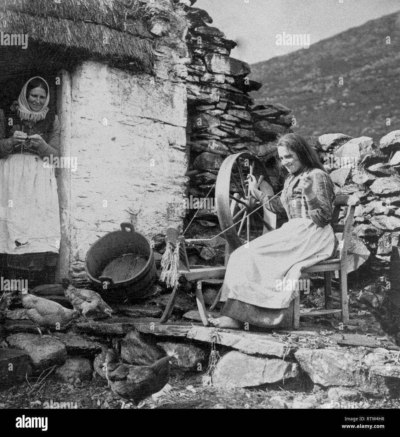 La parsimonia domestico vita reale in Irlanda 1904 Una donna seduta in corrispondenza di una ruota che gira al di fuori di una paglia-cottage con tetto in Irlanda rurale Foto Stock