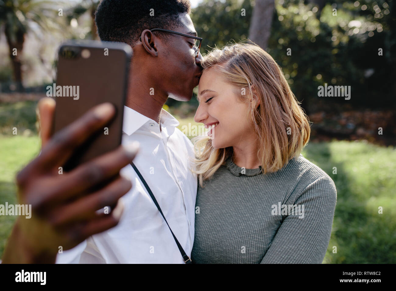 Amare giovane tenendo selfie con smart phone all'esterno. L'uomo baciare sulla fronte della sua fidanzata mentre sta assumendo il ritratto di auto. Foto Stock