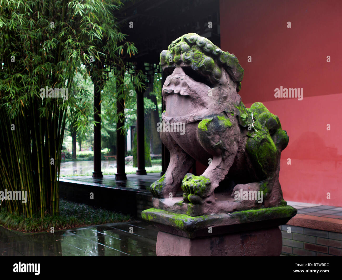 Mossy leone di pietra di fronte ad una costruzione Cinese con architettura tradizionale stile, accanto ad alberi di bambù. Foto Stock