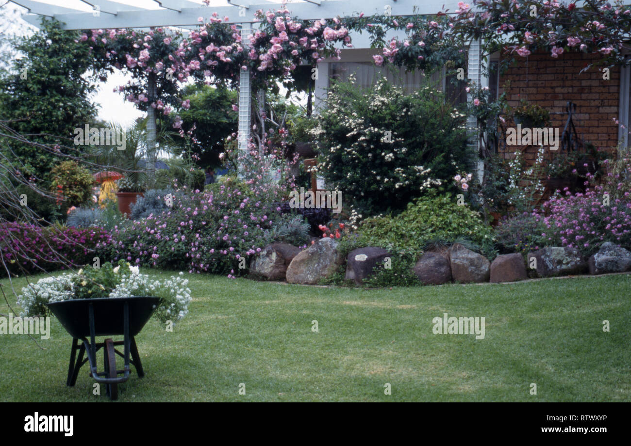 COUNTRY GARDEN con carriola, Prato e un assortimento di piante in giardino di roccia. Rosa Rambling Rosa pergola di copertura. Foto Stock