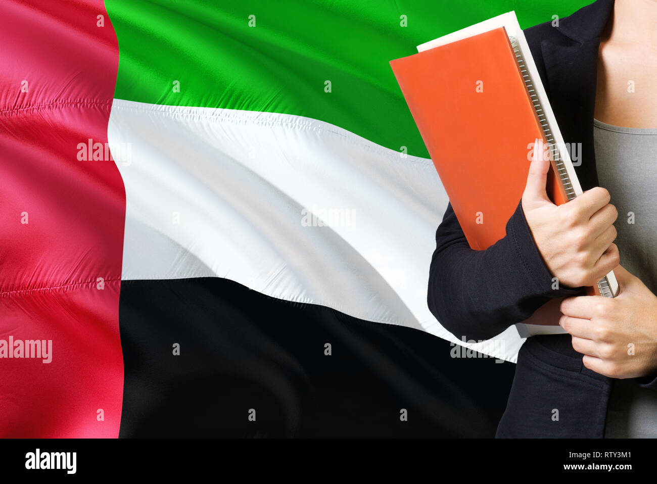 Apprendimento della lingua Emirian concetto. Giovane donna in piedi con la Bandiera degli Emirati Arabi Uniti in background. Docente in possesso di libri, arancione libro bianco Foto Stock