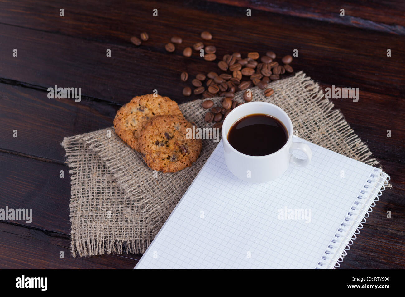 White tazza di caffè con caffè in chicchi vicino e biscotti e blocco note in una gabbia per i record su tessuto di lino sul vintage scuro dello sfondo. Foto Stock