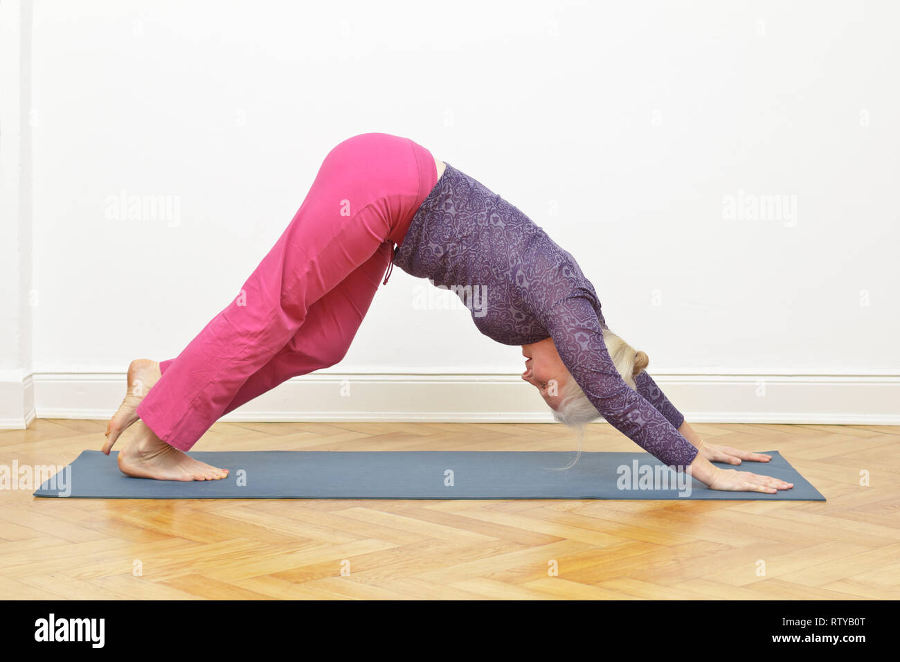 Senior donna esercizio di yoga in casa su un tappetino davanti a una parete bianca, asana cane a piedi Foto Stock