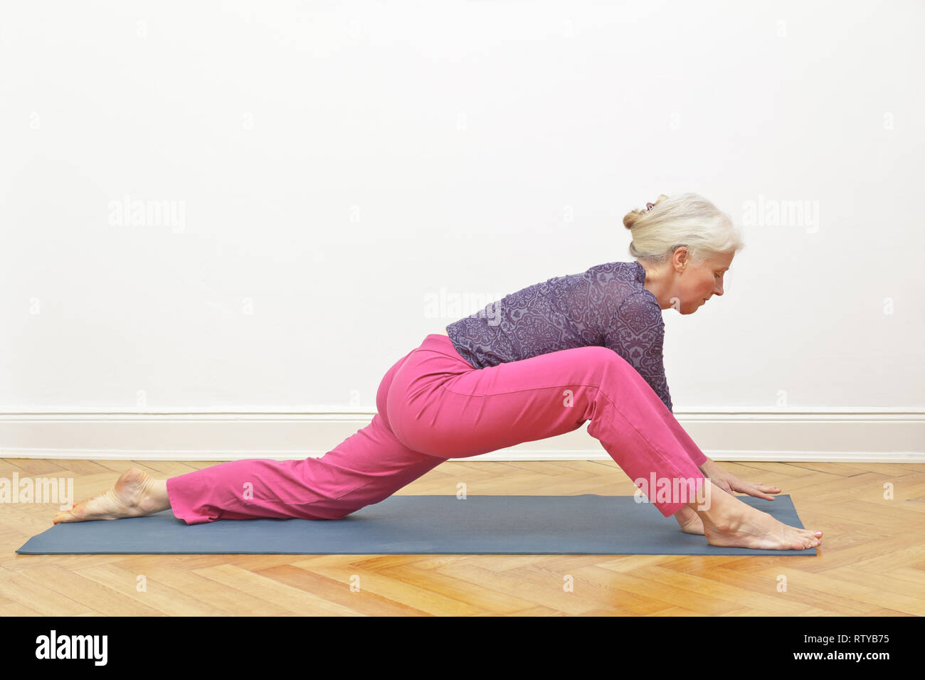 Senior donna con i capelli grigi facendo esercizi yoga a casa davanti ad un muro bianco, posizione dragon intro Foto Stock