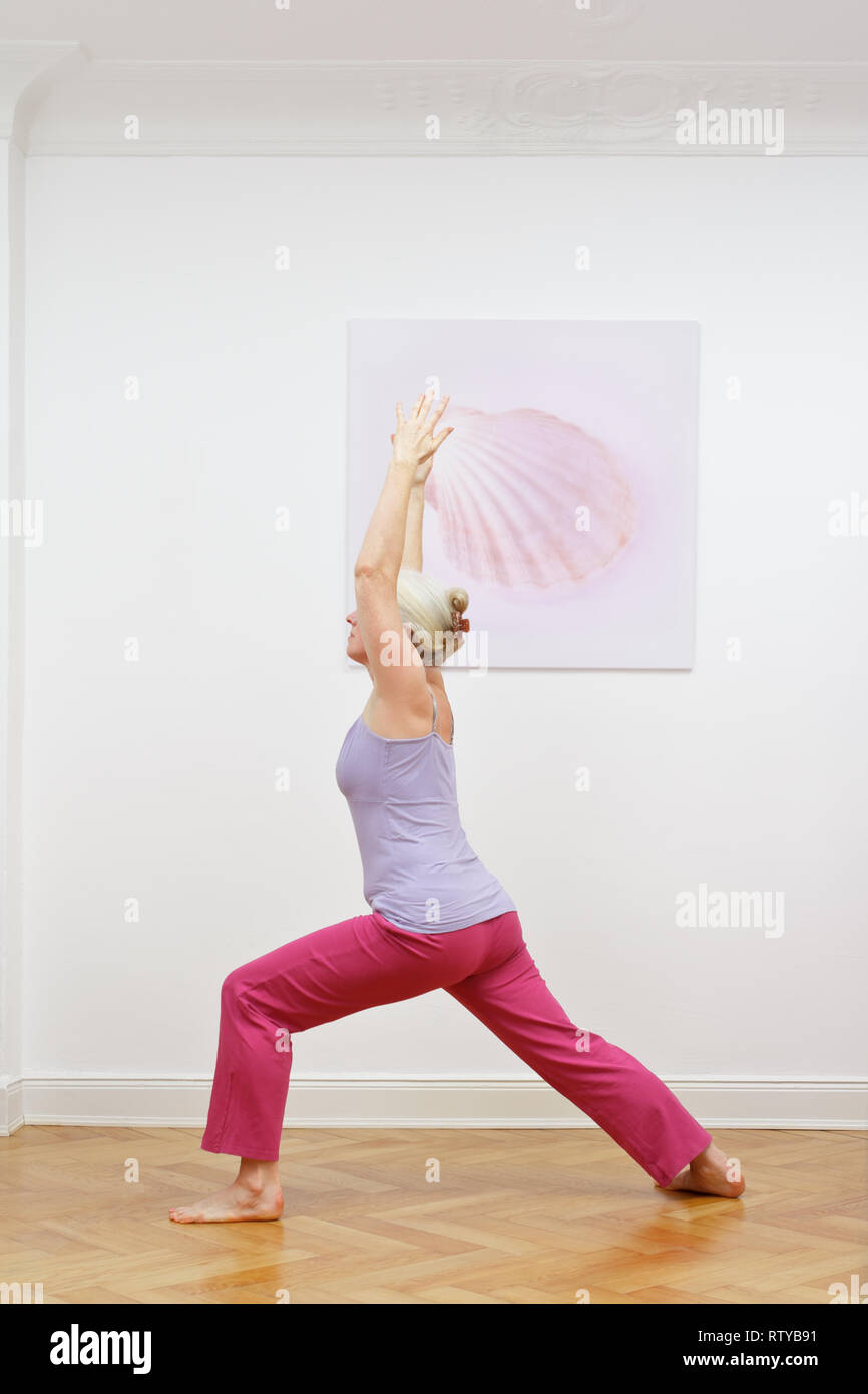 Senior donna con i capelli grigi facendo esercizi yoga a casa di fronte a un muro con una foto, asana GUERRIERO I Foto Stock