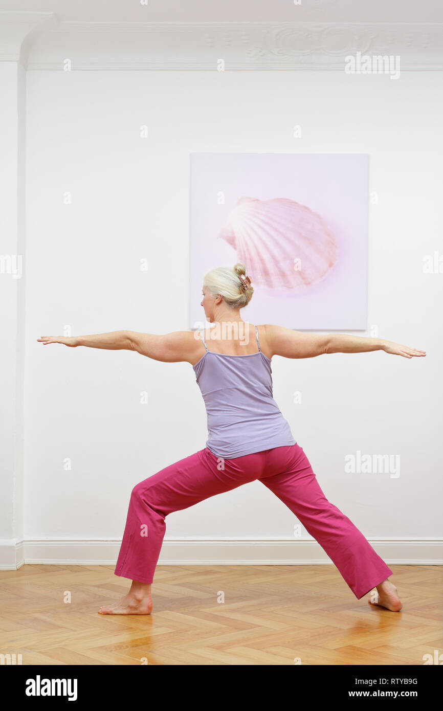 Senior donna con i capelli grigi facendo esercizi yoga a casa di fronte a un muro con una foto, asana GUERRIERO II Foto Stock