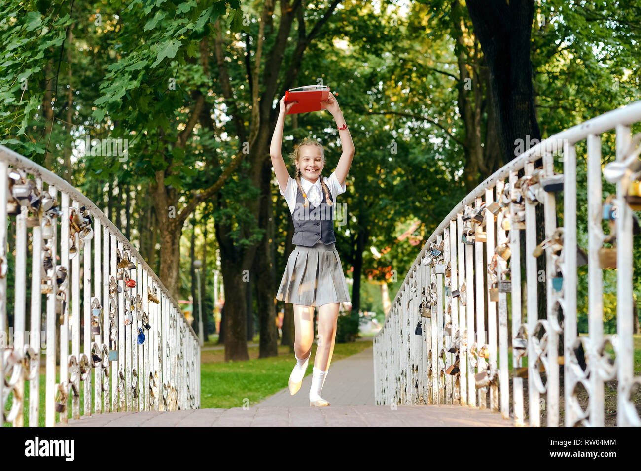 Studente schoolgirl felice con pig-tail in uniforme con libri in le mani sopra la testa scorre oltre il ponte in estate park Foto Stock