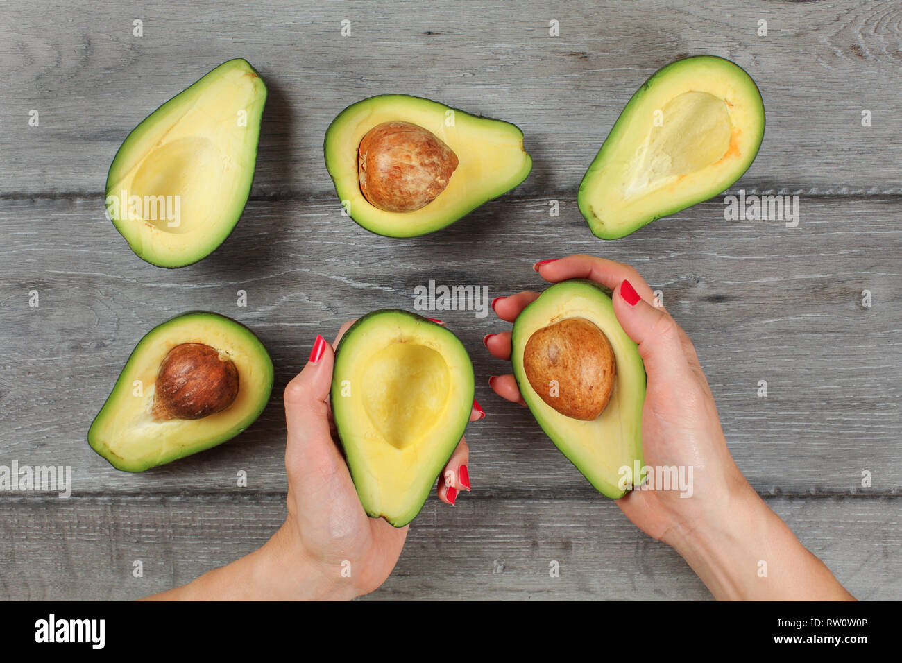 Vista del piano portapaziente - donna mani con unghie rosse, tenere gli avocadi tagliati a metà e metà più con sementi visibile su grigio scrivania di legno. Foto Stock