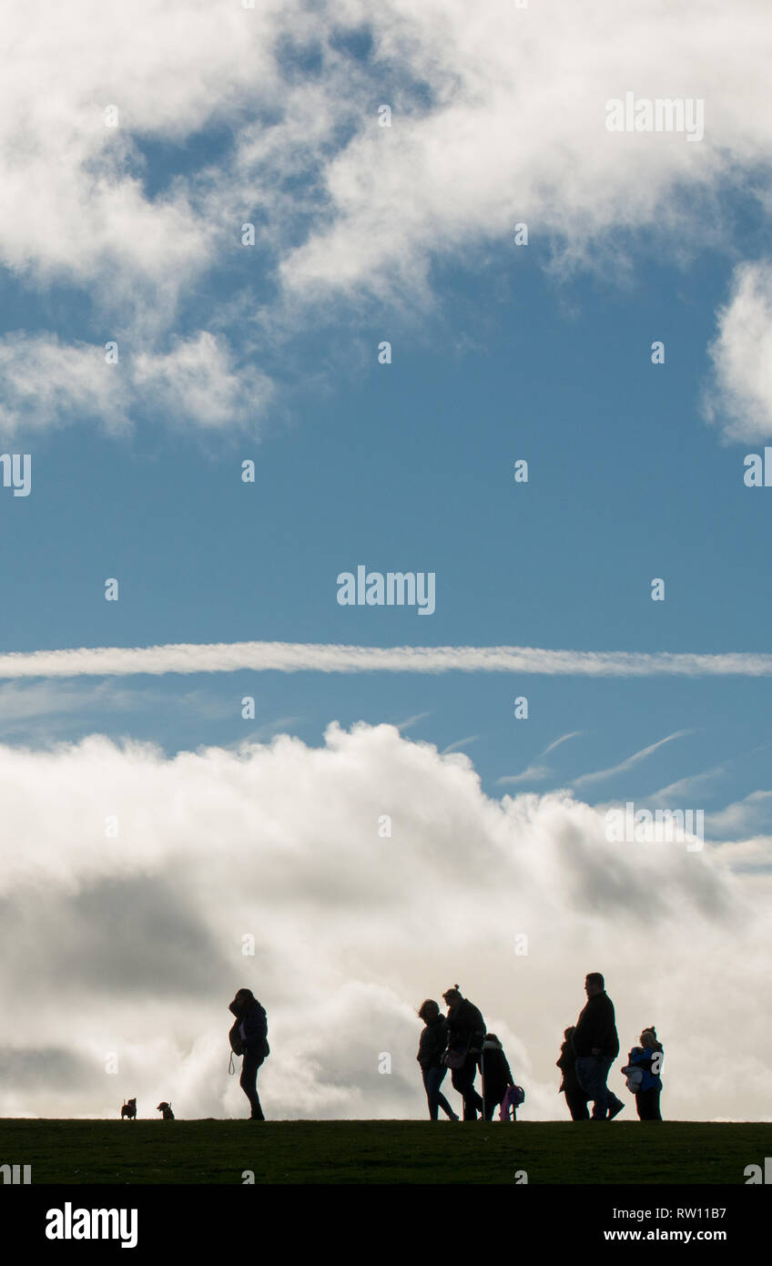 Un gruppo di persone con due cani fuori a piedi e catturati in silhouette contro una grande banca di nuvole e cielo blu profondo Foto Stock