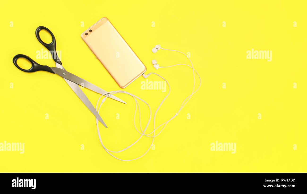 Vista dall'alto su rose gold telefono cellulare con auricolari cavo da tagliare con le forbici di grandi dimensioni su sfondo giallo. Foto Stock