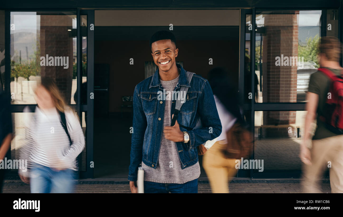 Ritratto di African ragazzo adolescente presso il college campus con studenti a piedi da in motion blur. Felice giovane ragazzo in piedi con il suo zaino guardando camer Foto Stock