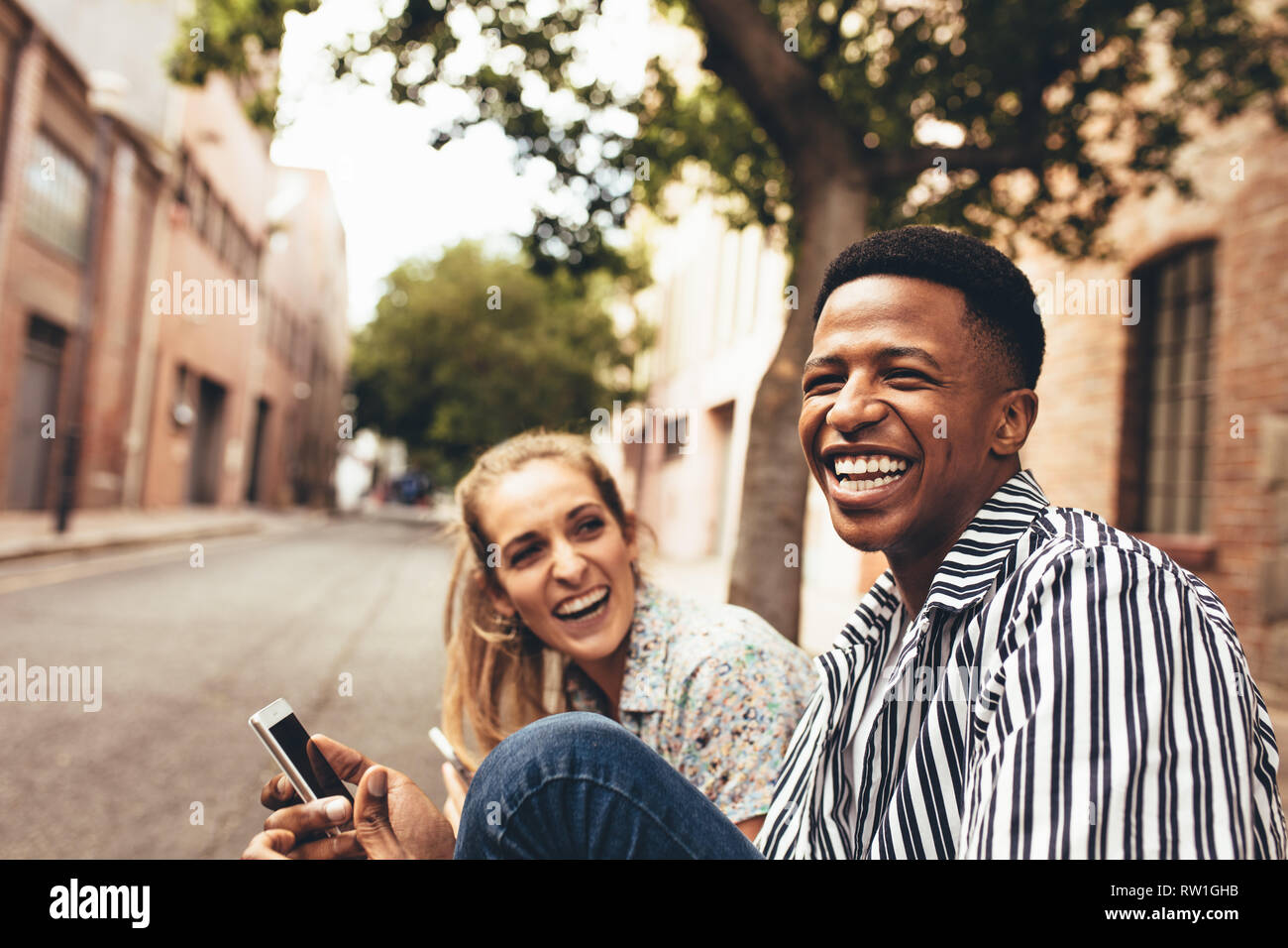Allegro multirazziale maschio e femmina amici seduti all'aperto con il telefono cellulare. Felice giovani amici all'esterno. Foto Stock