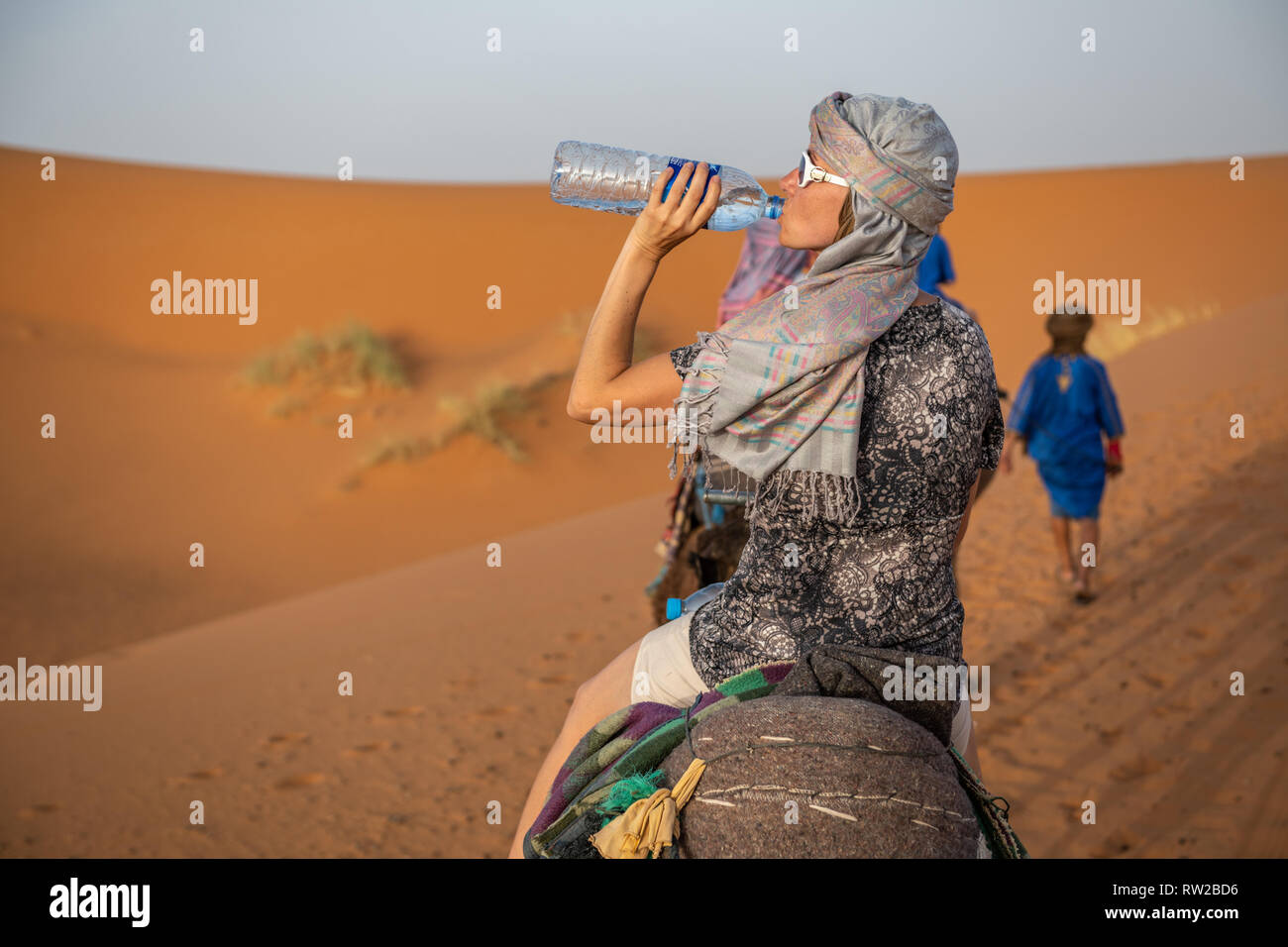 Una donna drink da un grande di acqua in bottiglia mentre a dorso di un cammello, Merzouga, Marocco Sahara Desert - Erg Chabbi dune Foto Stock