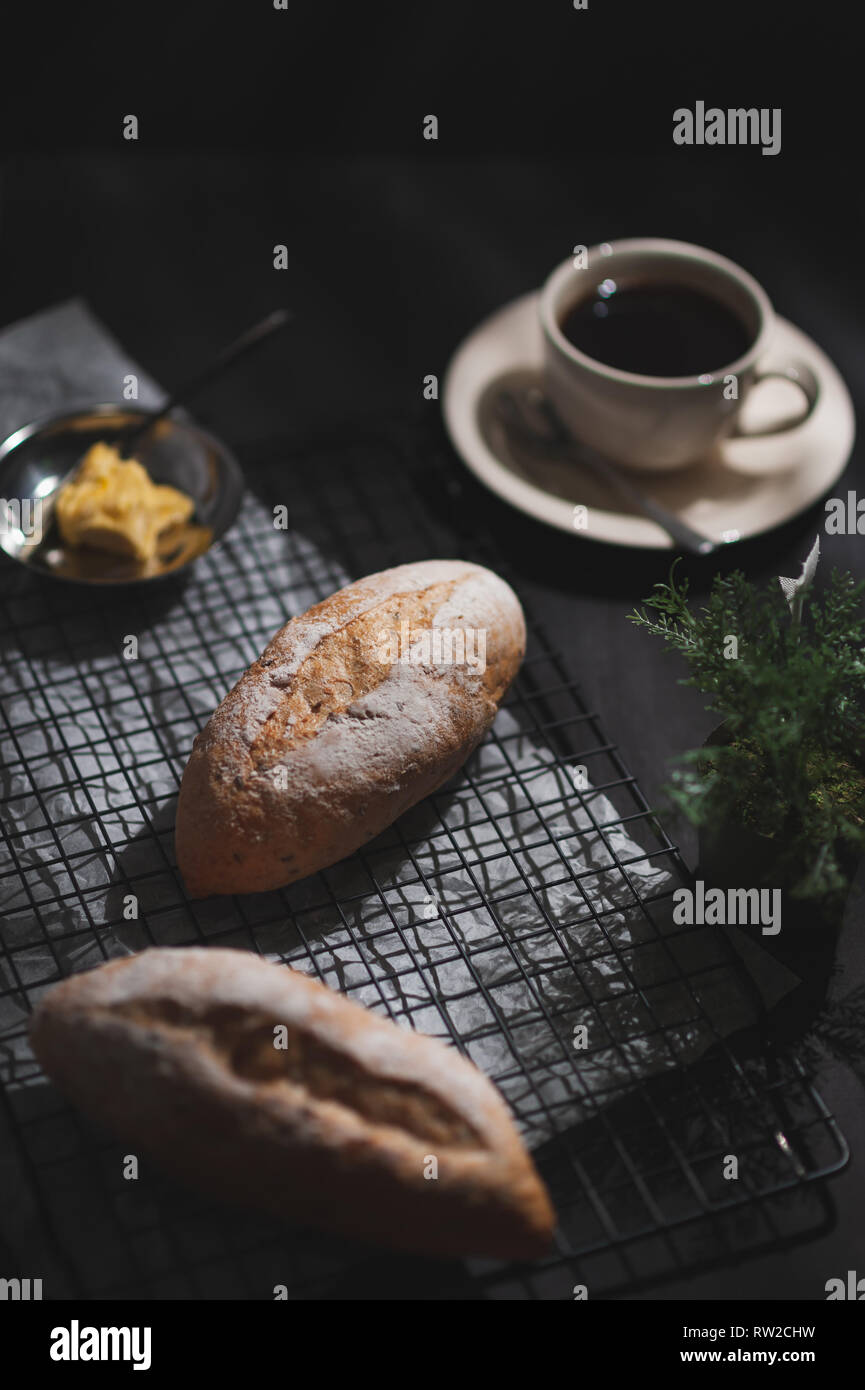 Baguette, in stile Europeo il pane con la margarina e il caffè nero su nero tavola in legno di prima mattina tempo duro con effetto di luce. Organici e salutari m Foto Stock