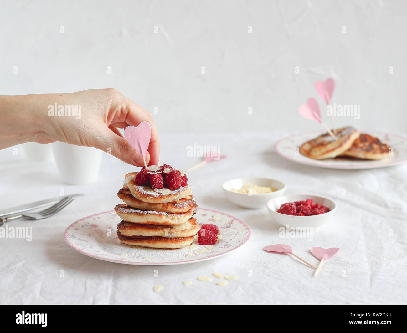 Tavolo per la colazione con una pila di frittelle, cuore decor e una mano collocando lamponi Foto Stock