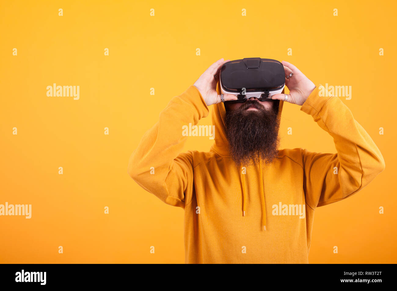 Hispter con barba lunga indossando felpa con cappuccio giallo e la realtà virtuale auricolare su sfondo giallo. Uomo bello. Barba lunga. Foto Stock