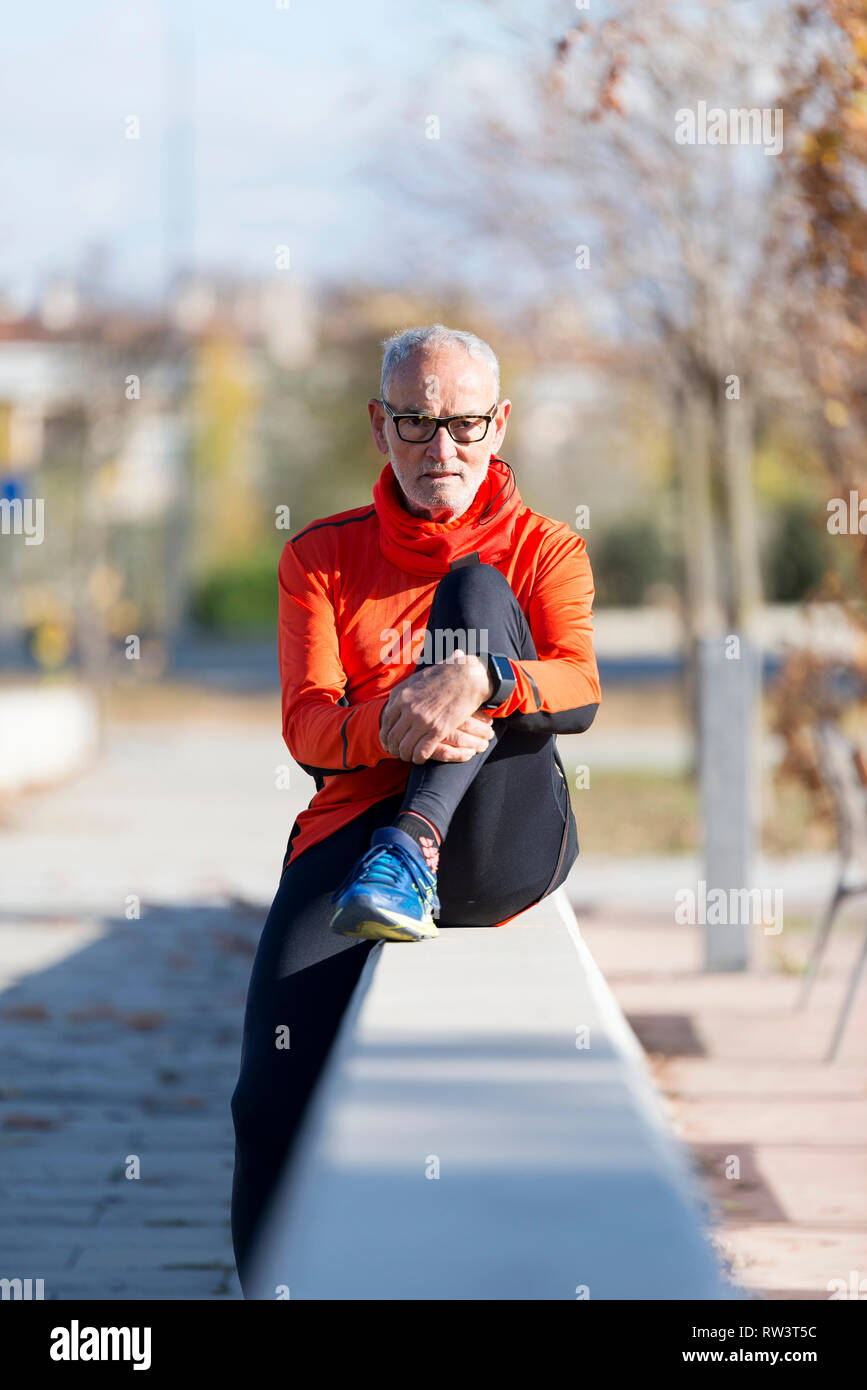 Vista frontale di un anziano uomo indossare abbigliamento e occhiali seduto su una recinzione rilassante guardando la fotocamera all'aperto in una giornata di sole Foto Stock