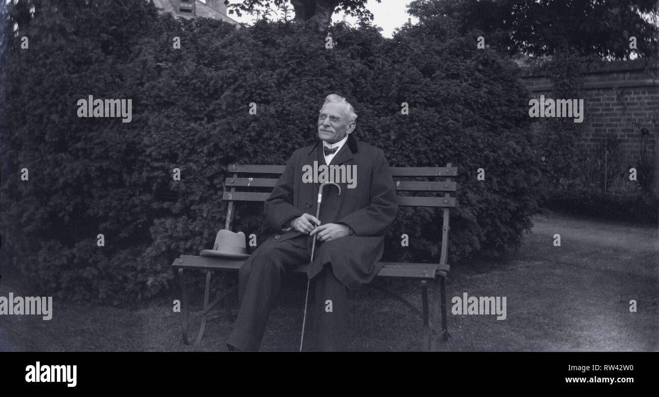 1930s, storico, anziano gentiluomo vestito in abiti formali del SER, indossando un bow tie e overcoast seduta su una panchina nel parco tenendo la sua canna da zucchero e con il suo cappello accanto a lui, Inghilterra, Regno Unito. Foto Stock