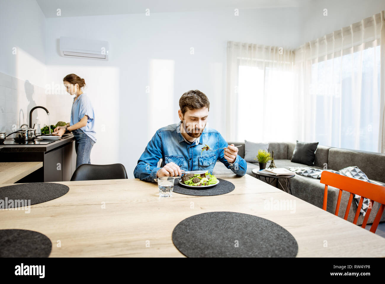 L'uomo mangiare insalata sana nel salotto del moderno appartamento con donna lavaggio piatti sullo sfondo Foto Stock