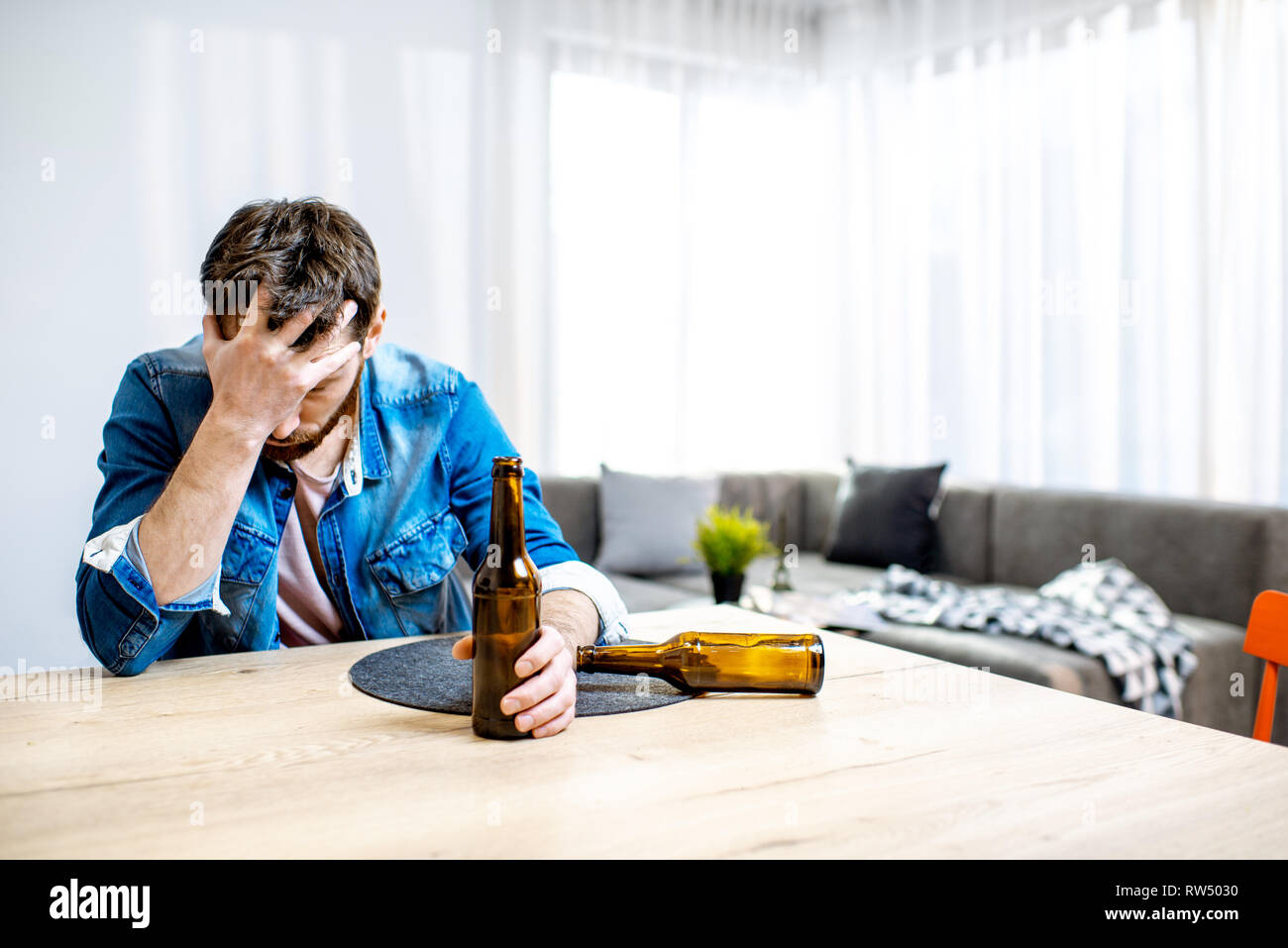 Bevuto alcolici maschile seduto con bottiglie sentirsi depressi e che soffrono di mal di testa a casa Foto Stock