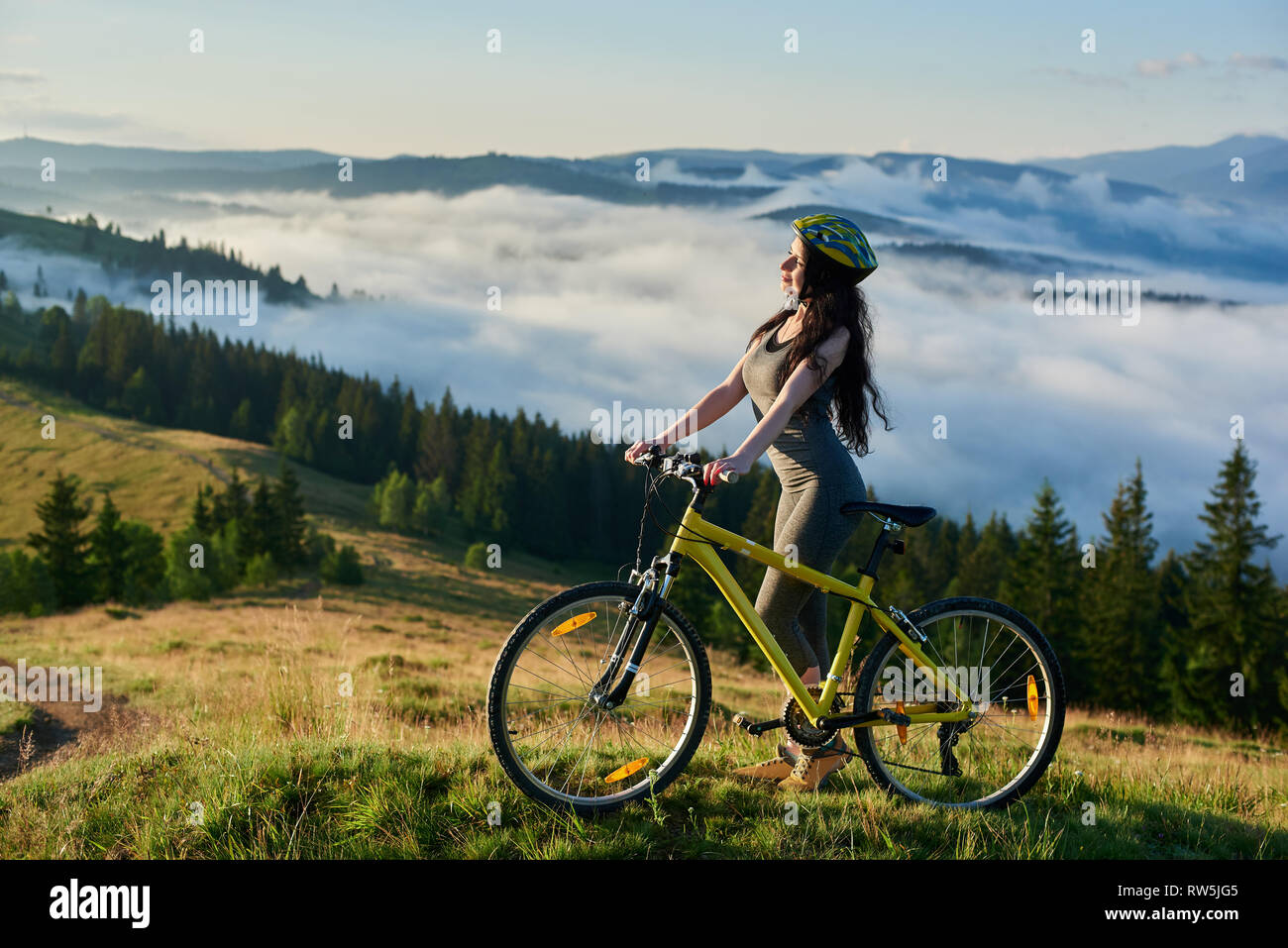 Femmina giovane conducente in piedi con giallo bicicletta in montagna, indossando il casco, godendo della vista della valle sulla mattina d'estate. Foggy montagne, foreste sullo sfondo sfocato. Il concetto di stile di vita Foto Stock