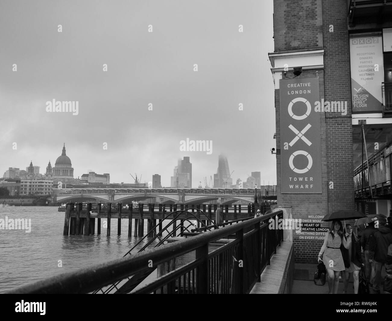 B&W immagine di due signore vestito per una notte fuori Londra, passando la OXO Tower sulla riva sud del Tamigi, ombrelli azienda contro la pioggia. Foto Stock