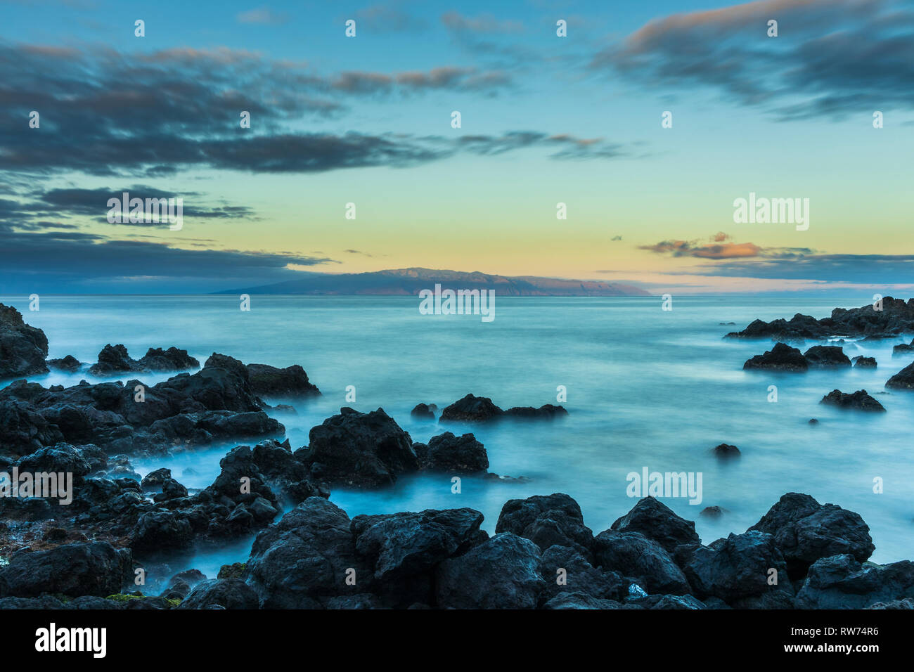 La Gomera all'orizzonte visto dalla Playa San Juan, Tenerife all'alba come il sole inizia appena a colpire l'isola, lunga esposizione con setosa movimento di acqua e rocce in primo piano, Isole Canarie, Spagna Foto Stock