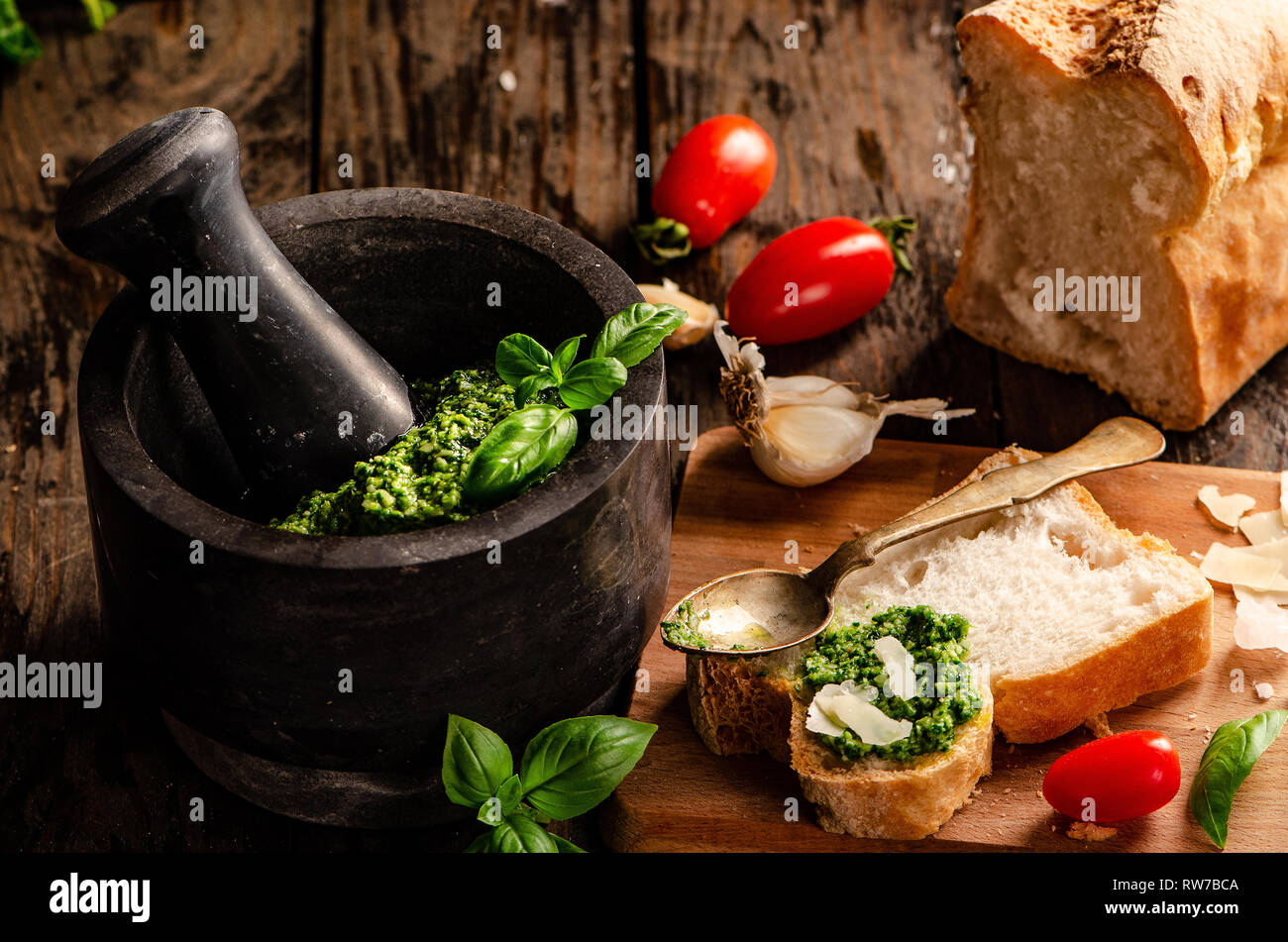 Mortaio con pesto genovese. Sfondo di legno con vari tipi di pane, basilico, aglio, pomodori e cucchiaio Foto Stock