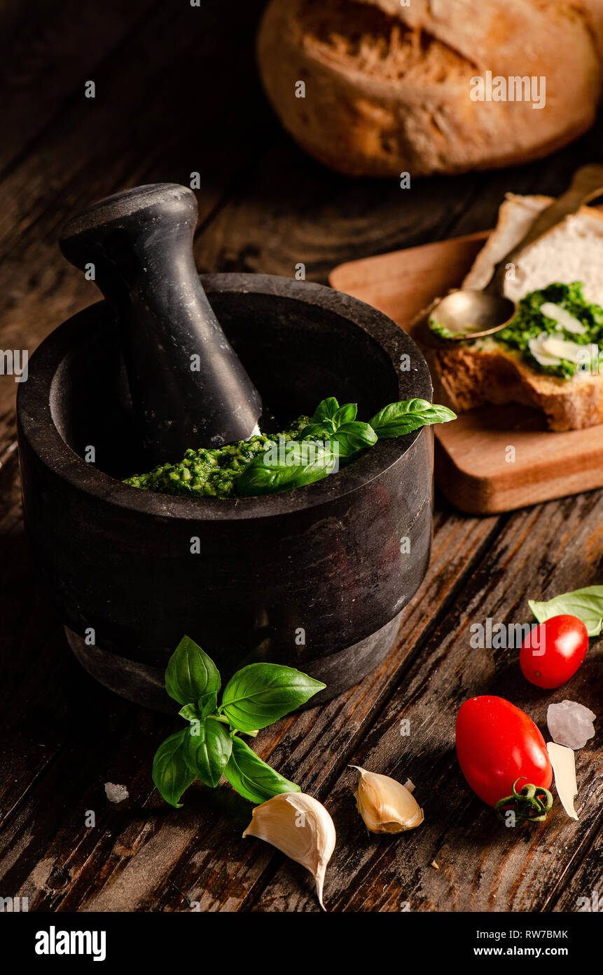 Mortaio con pesto genovese. Sfondo di legno con vari tipi di pane, basilico, aglio, pomodori e cucchiaio Foto Stock